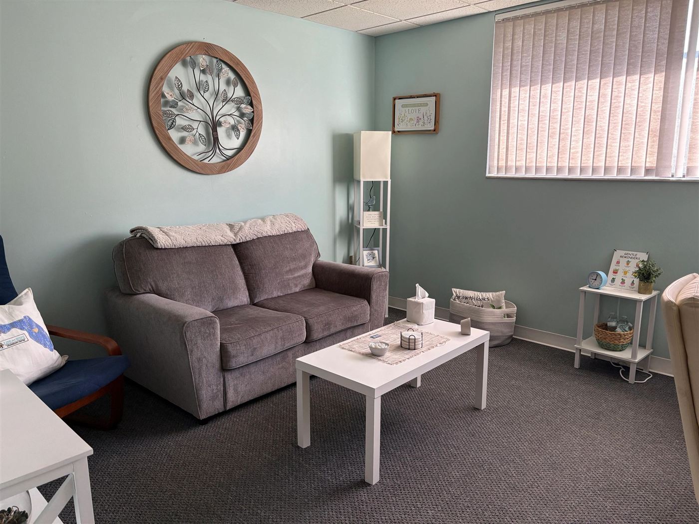 A counseling room with a grey loveseat, a round tree-of-life wall hanging and soft teal walls