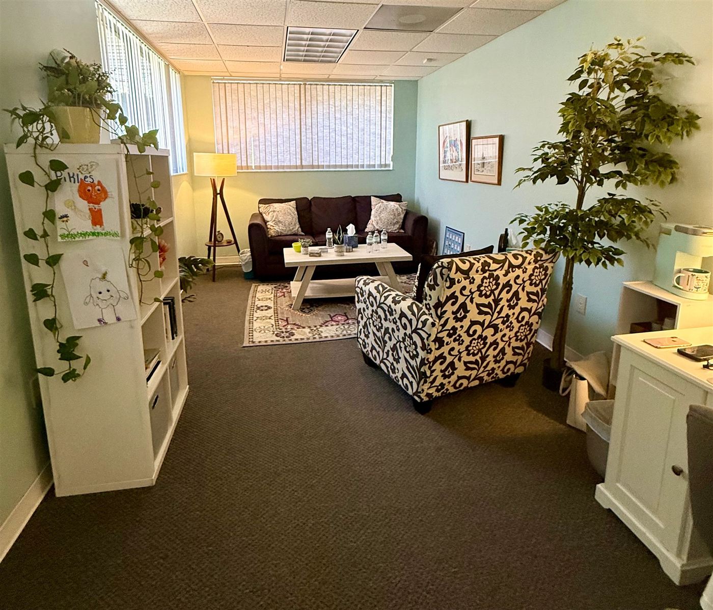 A warm counseling room with a sofa, a patterned armchair, plants, a floor lamp and children's drawings on a bookshelf